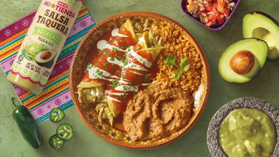 Mexican enchiladas topped with red sauce and sour cream, served with rice and refried beans, accompanied by H‑E‑B Mi Tienda Salsa Taquera Aguacate and a bowl of guacamole on a colorful tablecloth.