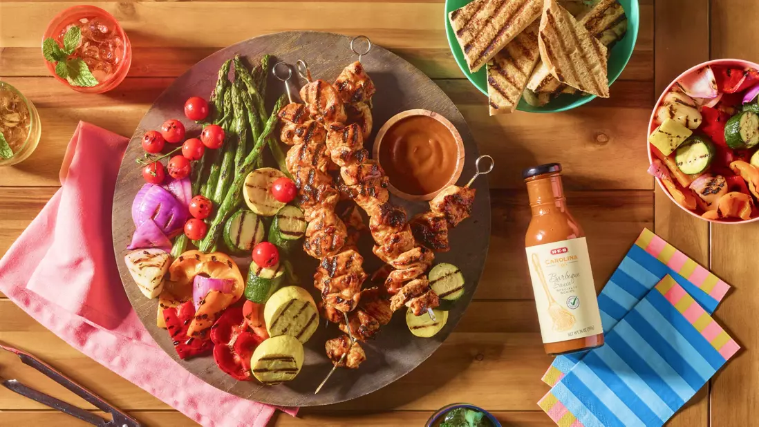 A summer meal loaded onto a plate with veggies in a bowl besides it and H‑E‑B carolina barbeque sauce next to it
