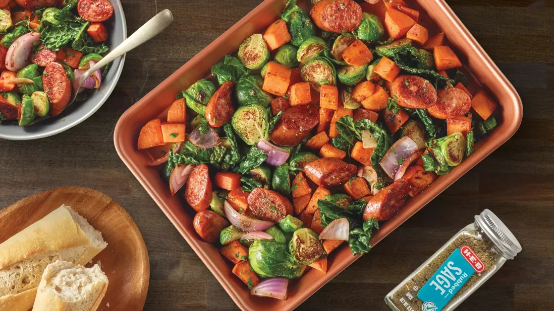 Sheet pan meal with roasted vegetables and sausage, including Brussels sprouts, sweet potatoes, red onions, and sausage slices, seasoned with sage. A side of bread and a bottle of H‑E‑B Rubbed Sage.