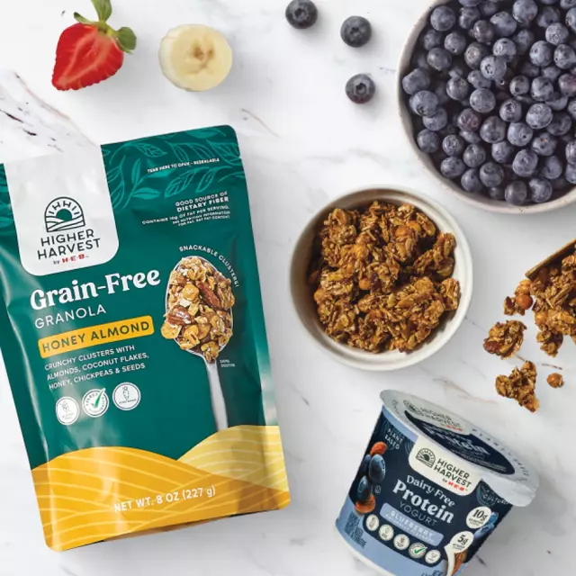 Higher Harvest by H‑E‑B Grain-Free Honey Almond Granola bag and bowl, Dairy-Free Blueberry Protein Yogurt cup, fresh blueberries, strawberries, and bananas on a marble surface.