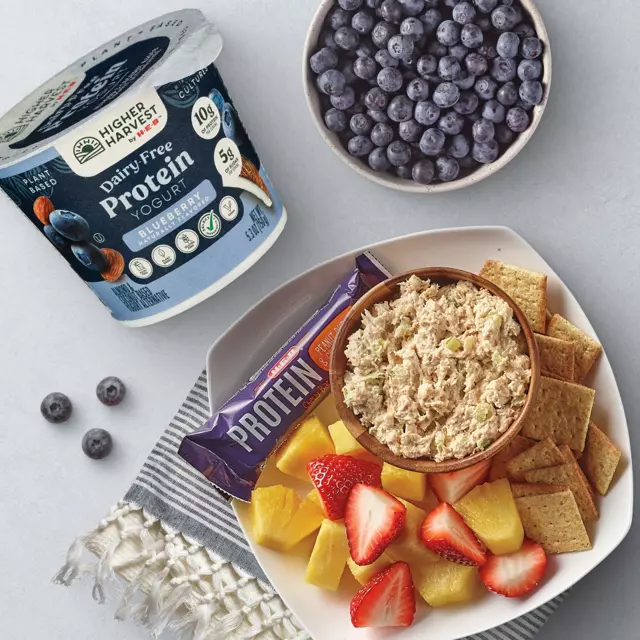 H‑E‑B Higher Harvest Dairy-Free Blueberry Protein Yogurt, fresh blueberries, chicken salad with crackers, pineapple, strawberries, and a protein bar on a table.