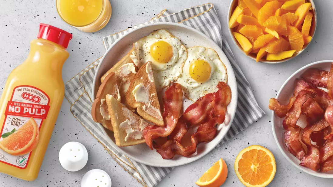 Juice (no pulp), toast, bacon and eggs on a plate with a bowl of fruit & bacon besides it