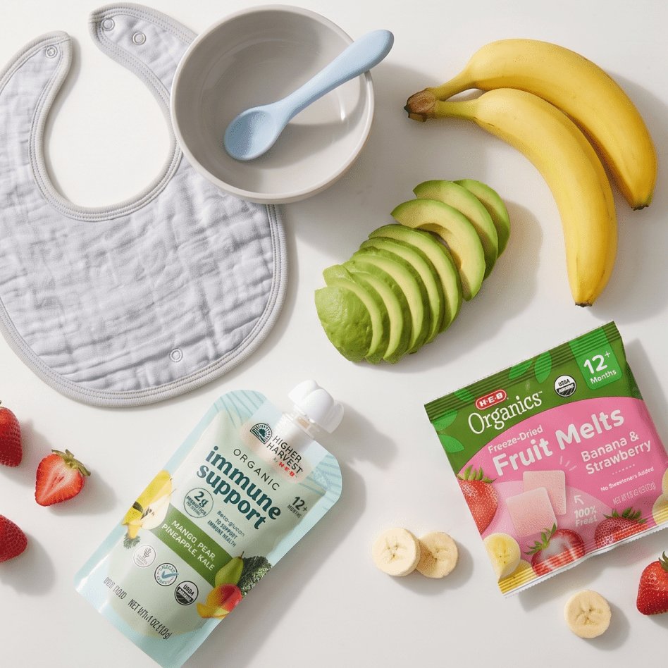 Flat lay of baby food essentials including a white bib, bowl with spoon, bananas, sliced avocado, strawberries, organic immune support puree, and freeze-dried fruit melts.
