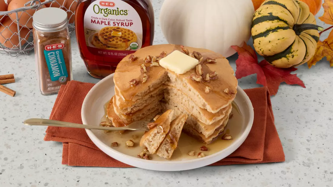 Banner featuring a stack of pumpkin pancakes topped with butter, maple syrup, and pecans, surrounded by fall decor including gourds and autumn leaves. H‑E‑B Organics maple syrup and pumpkin pie spice are displayed in the background.