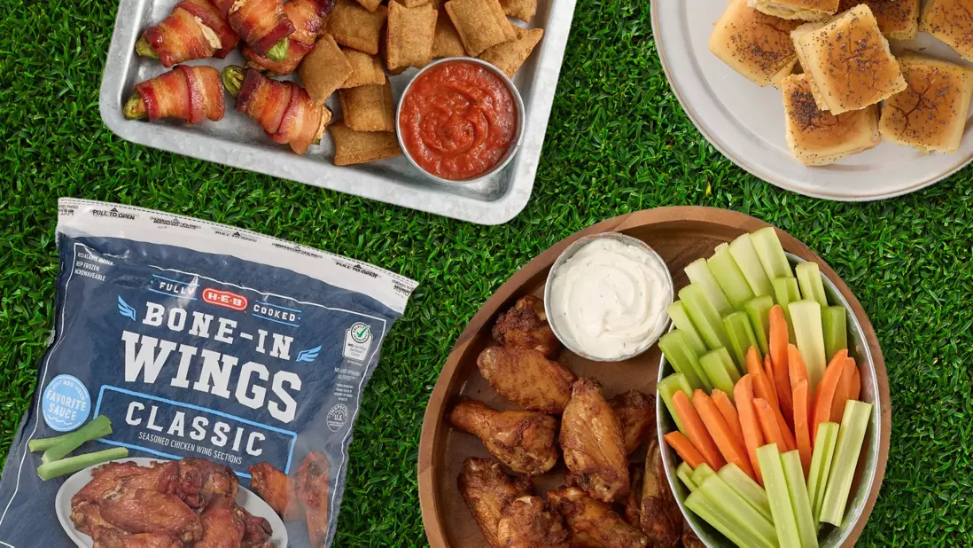 Game day snack spread featuring H‑E‑B fully cooked classic chicken wings, fresh celery and carrot sticks with dipping sauce, bacon-wrapped appetizers, pizza rolls, and garlic bread on a football field-themed background.