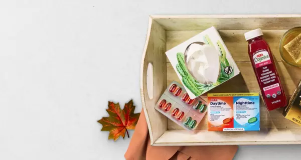 Cold and flu relief promotional banner featuring the headline “Relief for the sniffles” on a light gray background. A wooden tray on the right contains wellness and cold-care essentials, including facial tissues, throat lozenges, DayQuil and NyQuil medications, an immune support beverage, herbal tea, and honey. A fall leaf accent appears near the bottom, reinforcing a seasonal wellness theme.