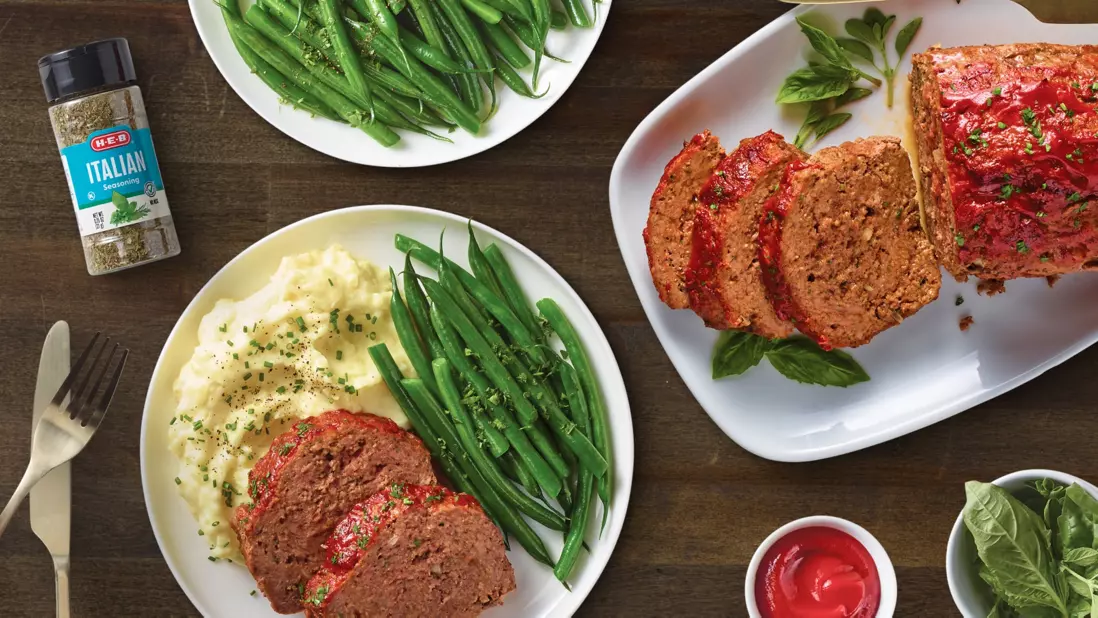 Prepared meatloaf dinner served with mashed potatoes and green beans alongside sliced meatloaf on a serving platter. An H‑E‑B Italian seasoning bottle appears nearby, with fresh basil leaves and ketchup completing the hearty meal presentation.