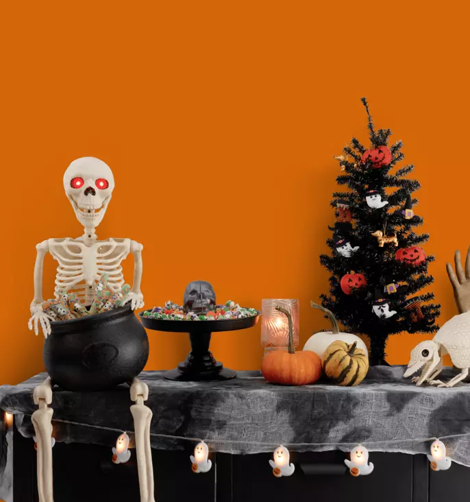 Halloween décor display featuring a skeleton seated beside a cauldron of candy, a black Halloween tree with pumpkin ornaments, pumpkins, ghost string lights, and spooky decorations. An orange background highlights the message “Spooky season is here.”