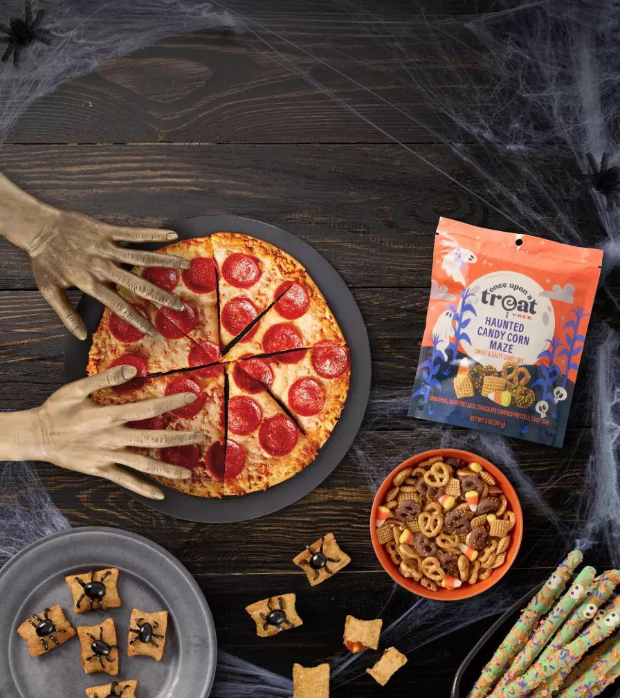 Halloween-themed snack spread on a dark wood table with cobweb decorations. Pepperoni pizza topped with spooky hand props, candy corn mix, decorated pretzel sticks, spider-themed crackers, and assorted treats surround the display.