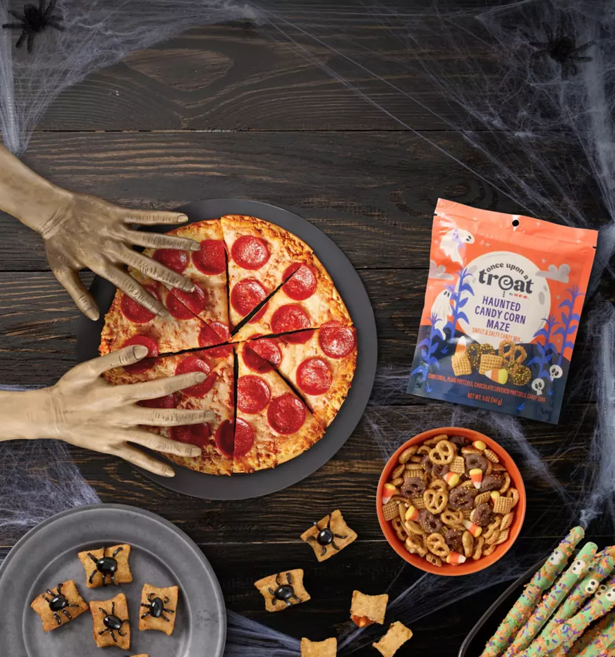 Halloween party food spread on a dark wood table with pepperoni pizza, decorated cookie bites, pretzels, candy corn, and Halloween-themed snacks. Cobweb decorations and spooky hands reaching toward the pizza accompany the headline “Scary-good party musts.”