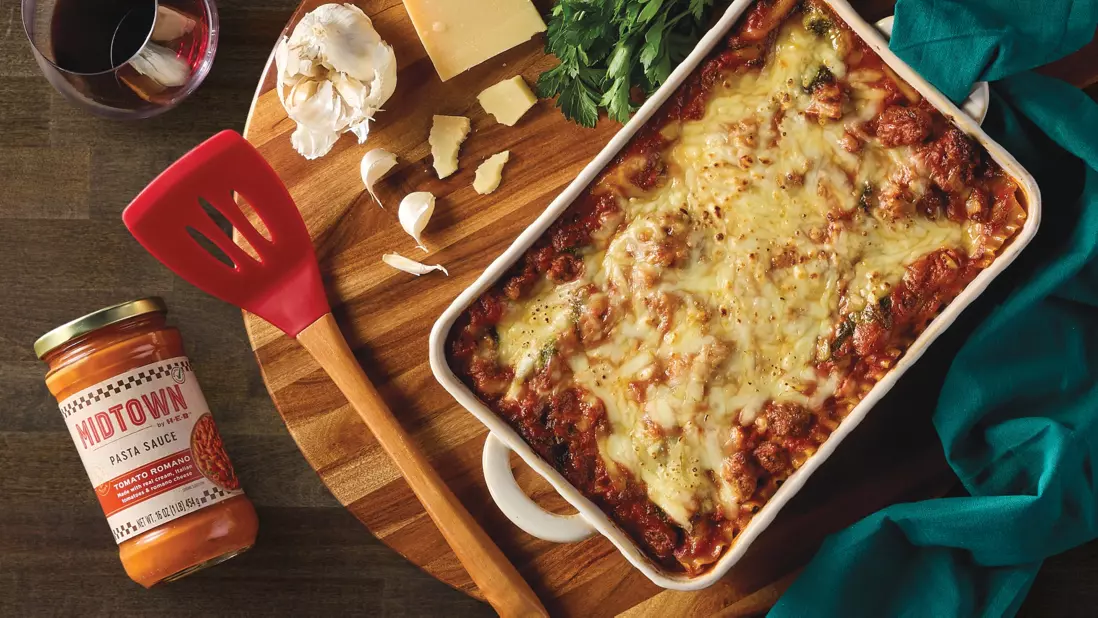 Baked lasagna topped with melted cheese in a white casserole dish on a wooden board. Surrounding ingredients include a jar of Midtown pasta sauce, garlic cloves, cheese wedges, parsley, a red spatula, and a glass of red wine.