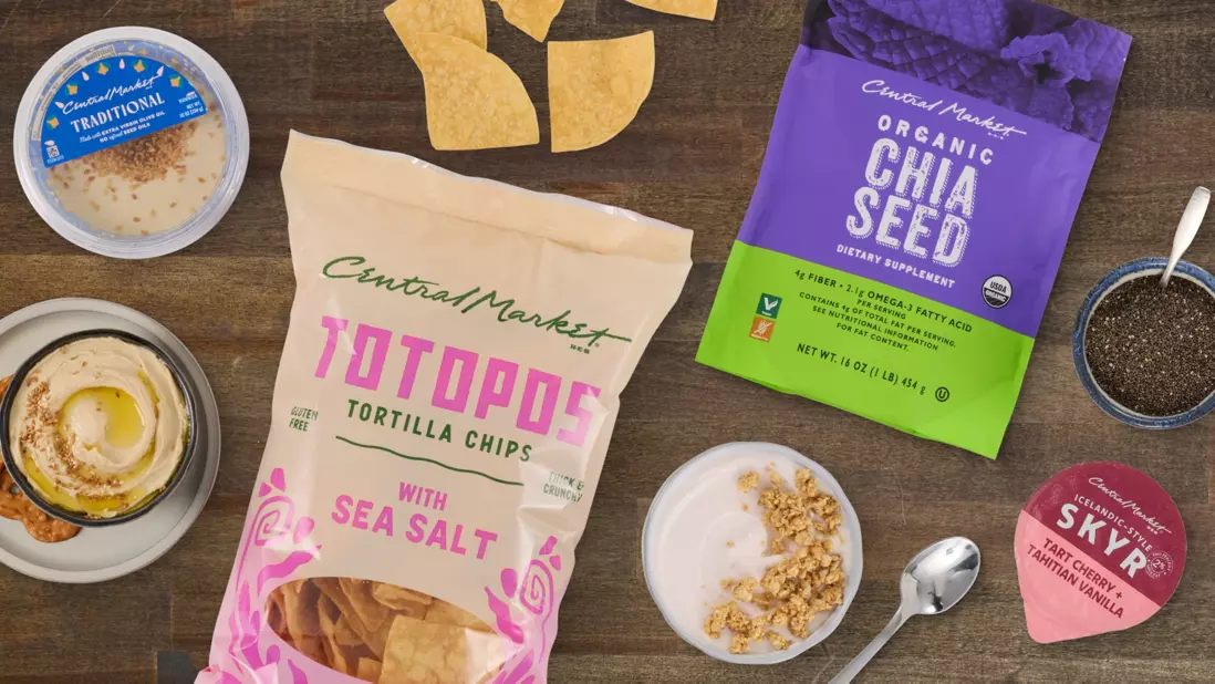 Assortment of snack and breakfast items on a wooden surface, including Central Market sea salt tortilla chips, organic chia seeds, traditional hummus, skyr yogurt, tortilla chips, chia seeds in a bowl, and granola.