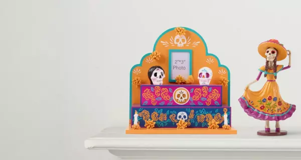 Banner featuring a colorful Day of the Dead altar display with decorative skulls, flowers, candles, and framed artwork. A festive Day of the Dead figurine stands beside the display. Text reads “Celebrate Day of the Dead.”