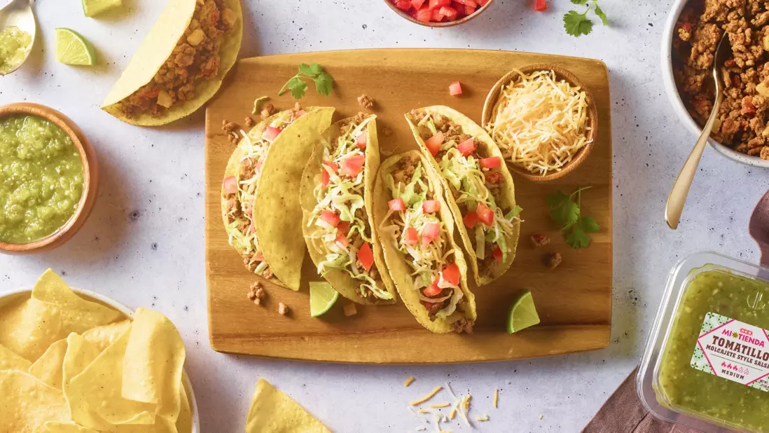 Banner featuring three crispy tacos topped with seasoned meat, shredded lettuce, diced tomatoes, and shredded cheese on a wooden board. Surrounded by tortilla chips, salsa verde, shredded cheese, taco meat, lime wedges, and tomatillo salsa.
