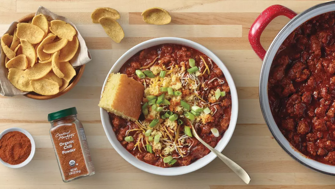 Banner featuring a bowl of chili topped with shredded cheese and green onions, served with cornbread. Surrounding items include tortilla chips, chili seasoning, and a pot of chili. The meal is displayed on a wooden countertop.