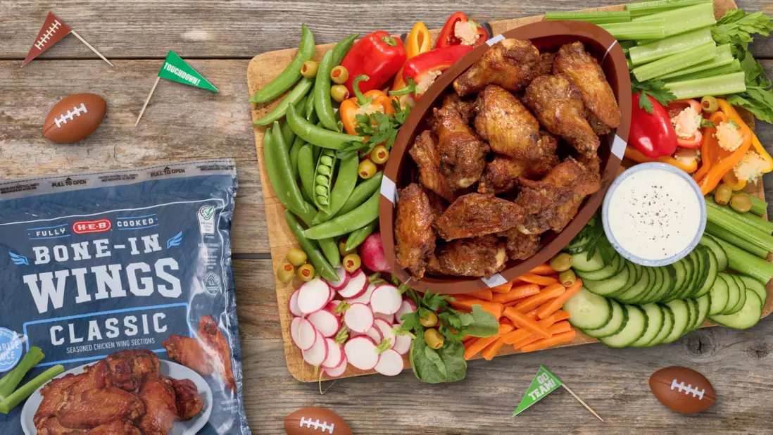 H‑E‑B Cooked Bone-In Wings served in a football-shaped bowl with celery, cucumbers, carrots, radishes, snap peas, peppers, and dipping sauce. A package of H‑E‑B Bone-In Wings and football-themed decorations complete the game day spread.