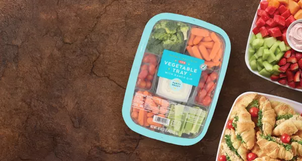 Get $2 off select party trays. Image features a fruit tray with strawberries, grapes, melon, pineapple, and dip, a tray of croissant sandwiches with tomatoes, and a vegetable tray with ranch dip on a rustic tabletop.