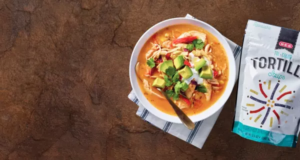 Feel-good dinners for tonight. A bowl of creamy tortilla soup topped with avocado, cilantro, sour cream, and tortilla strips sits beside a bag of H‑E‑B tri-color tortilla strips on a rustic brown surface.