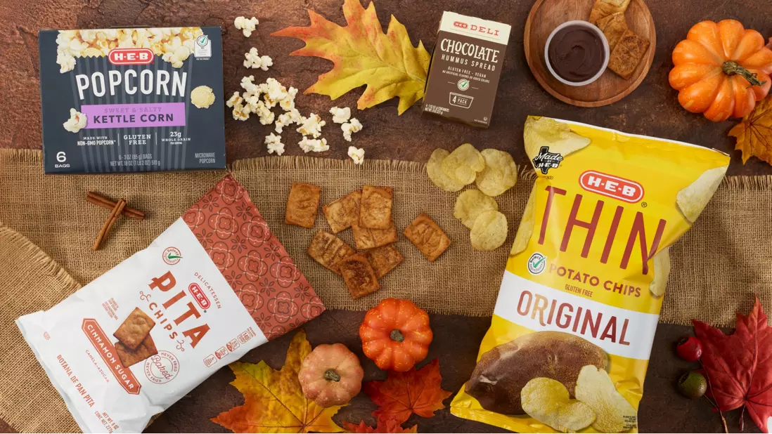 Fall snack spread featuring H‑E‑B Thin Original Potato Chips, H‑E‑B Chicharrones, H‑E‑B Sweet & Salty Kettle Corn Popcorn, chocolate brownie bites with dip, crackers, and autumn leaves and mini pumpkins on a rustic tabletop.