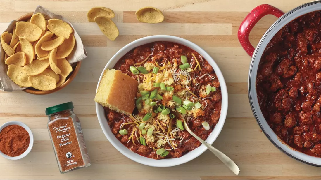 Bowl of hearty chili topped with shredded cheese, green onions, and crispy tortilla strips, served with cornbread. A slow cooker filled with chili, tortilla chips, and H‑E‑B chili seasoning complete the meal on a wooden tabletop.