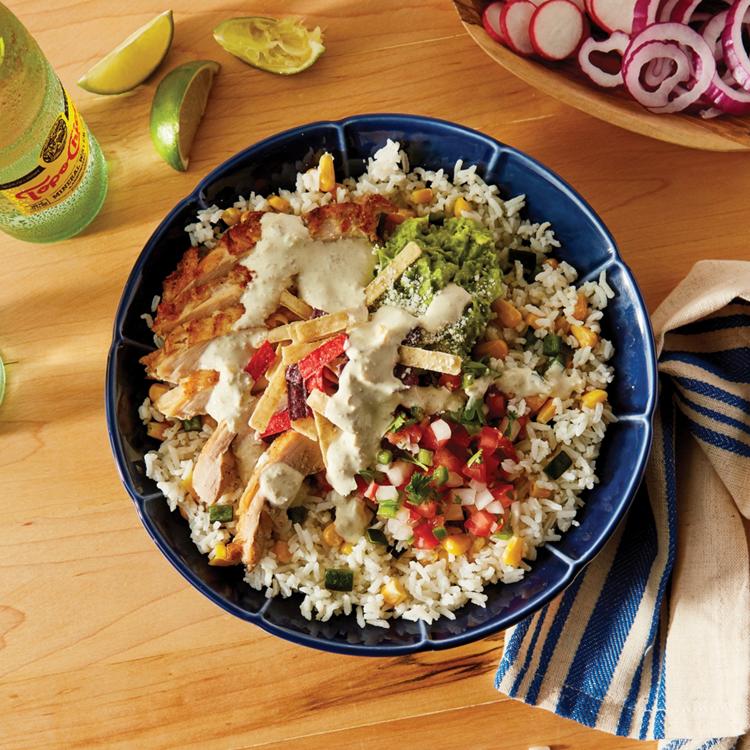 Grilled fajita chicken bowl on corn and poblano white rice and topped with guacamole, cotija cheese, pico de gallo, and tortilla strips. The fajita bowl is on a table with lime wedges on the side and a topo chico.