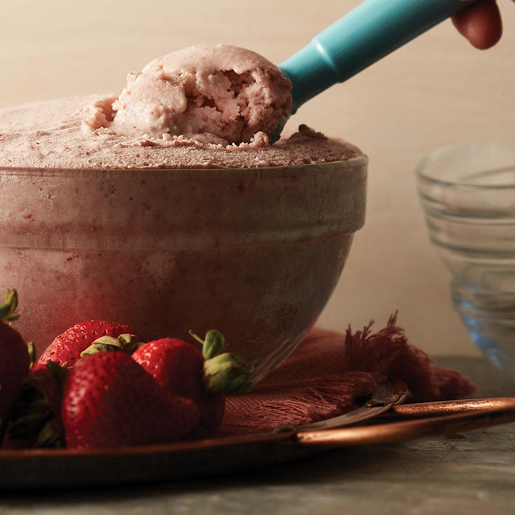 An ice cream scoop dipping into a glass bowl of homemade ice cream with strawberries on the side