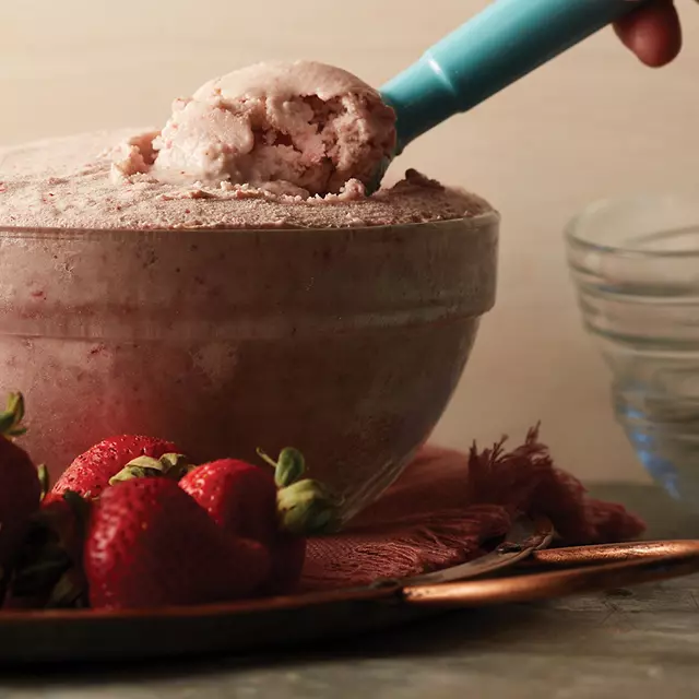 An ice cream scoop dipping into a glass bowl of homemade ice cream with strawberries on the side