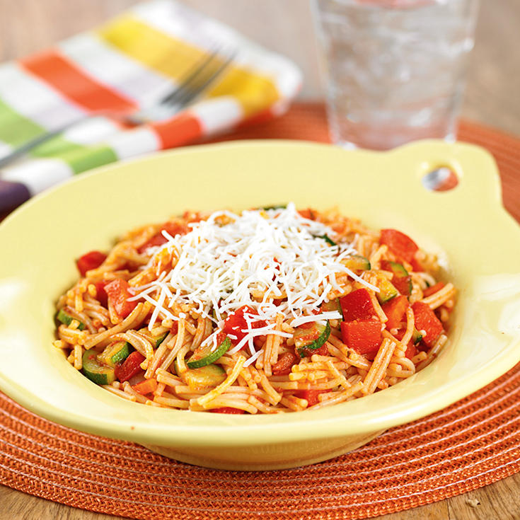 A bowl of cut spaghetti noodles with small bites of zucchini and other vegetables, topped with shredded cheese