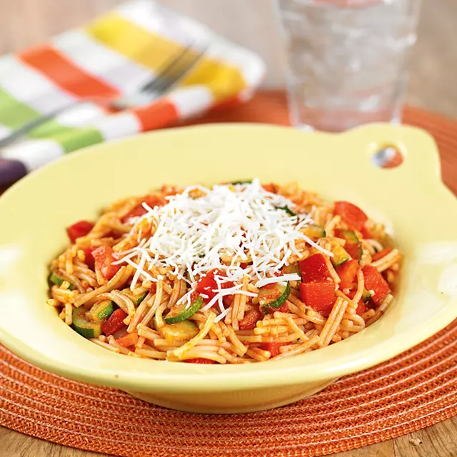 A bowl of cut spaghetti noodles with small bites of zucchini and other vegetables, topped with shredded cheese
